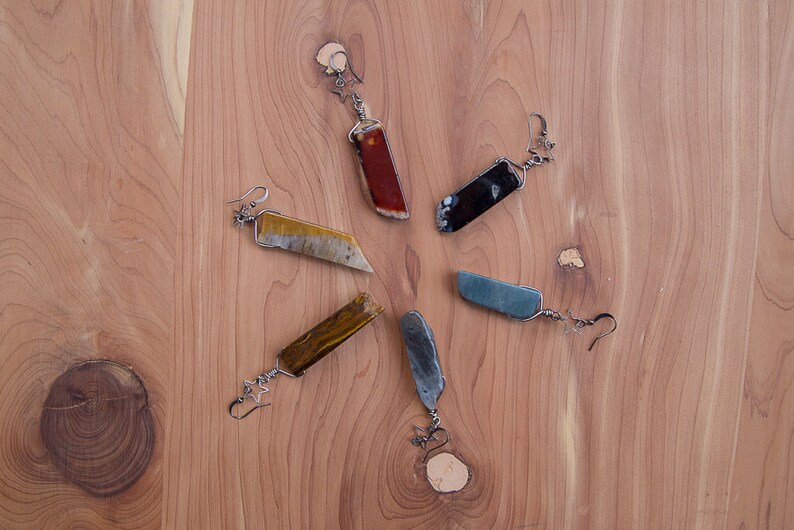 May include: Six pairs of earrings with different coloured stone accents. The earrings are made of metal and have a simple design. The stones are all rectangular in shape and are set in a silver tone metal.