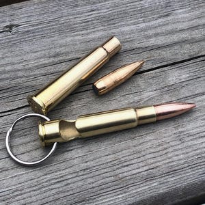 May include: A brass bottle opener shaped like a rifle cartridge with a silver metal keyring attached to the base. Beside it lies a separate brass cartridge casing and a copper-coloured bullet tip, all featuring a polished metallic finish.
