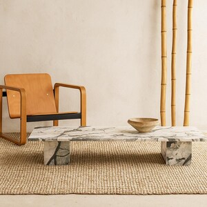 May include: A rectangular marble coffee table with gray veining, supported by two square marble bases. A wooden bowl sits on the table. A light brown wooden chair and bamboo stalks are in the background. The table is on a woven rug.