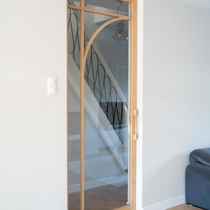 Op de afbeelding: Een glazen deur met een houten frame en een decoratief zwart metalen ontwerp. De deur heeft een lange houten handgreep en is geplaatst in een witte muur. Een trap is zichtbaar door het glas en een grijze bank staat op de achtergrond.