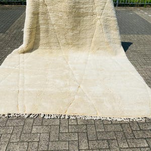 May include: A large, cream-coloured rug with a geometric diamond pattern. The rug has a thick, textured pile and a fringe along the bottom edge. The rug is displayed outdoors on a brick surface.