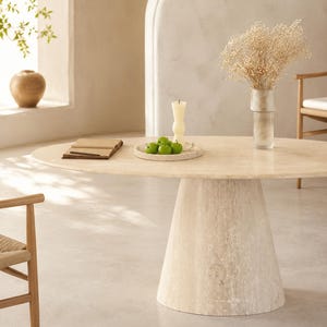 May include: An oval dining table in light beige, supported by a conical base. The table is adorned with green apples on a plate, a candle, and a vase of dried flowers. A wooden chair is in view.