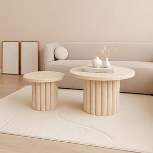 May include: Two beige, cylindrical coffee tables with round tops. The larger table has a book and vases on top. The tables are on a cream-colored rug in front of a beige sofa. The room has a minimalist design.