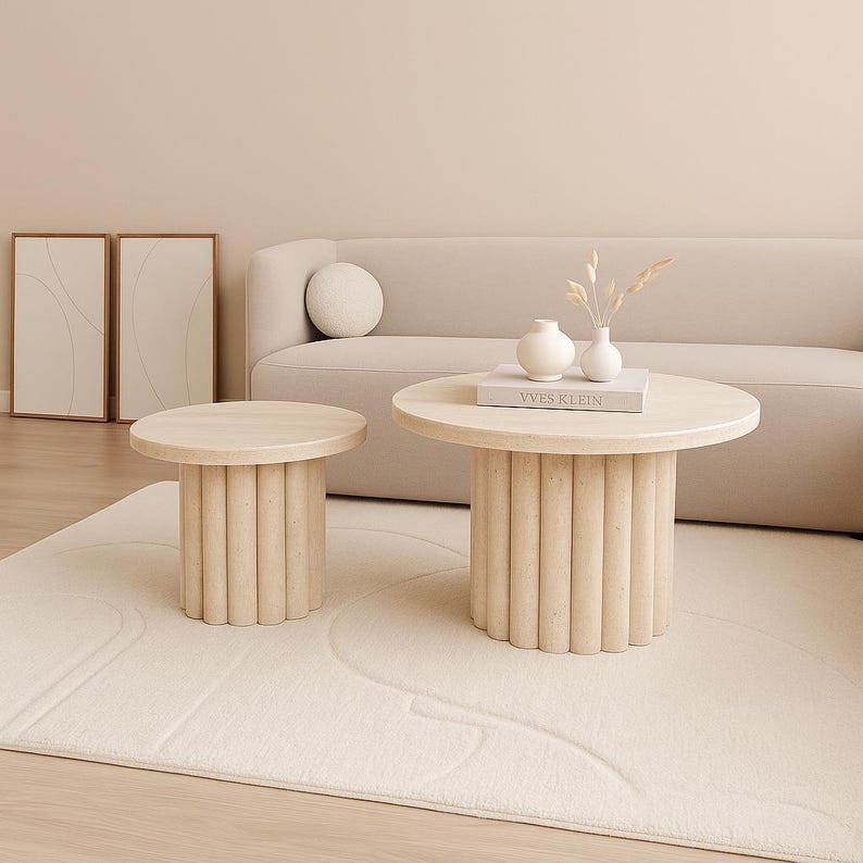 May include: Two beige, cylindrical coffee tables with round tops. The larger table has a book and vases on top. The tables are on a cream-colored rug in front of a beige sofa. The room has a minimalist design.