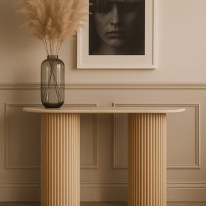 Könnte beinhalten: Eine moderne Konsole aus hellem Holz mit zwei zylindrischen, geriffelten Beinen. Ein gerahmtes Schwarz-Weiß-Porträt hängt darüber. Eine hohe, graue Glasvase enthält getrocknetes Pampasgras.