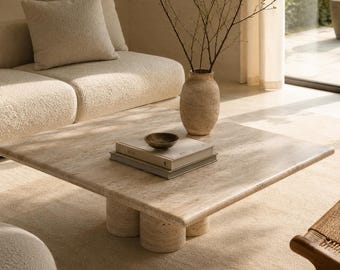 May include: A square, beige travertine coffee table with rounded cylindrical legs. The table holds a stack of books, a small bowl, and a textured vase with branches. A beige sofa and rug complete the neutral-toned living room.