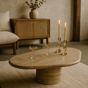 Könnte beinhalten: Ein hellbrauner, ovaler Couchtisch mit zylindrischem Sockel. Zwei brennende Kerzen in goldfarbenen Haltern und zwei Champagnergläser stehen auf dem Tisch. Ein beiger Teppich und ein Holzschrank befinden sich im Hintergrund.