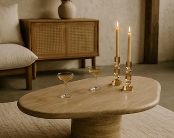 May include: A light brown, oval-shaped coffee table with a cylindrical base. Two lit candles in gold-colored holders and two champagne glasses sit on the table. A beige rug and a wooden cabinet are in the background.