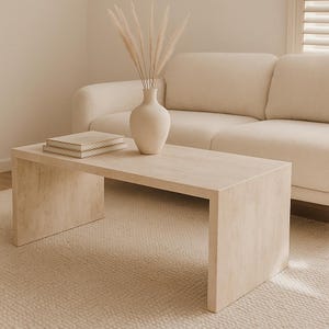 Op de afbeelding: Een rechthoekige, lichtbeige salontafel met een minimalistisch design. Een stapel boeken en een crèmekleurige vaas met gedroogde planten staan op de tafel. Op de achtergrond staat een crèmekleurige bank. De tafel is ongeveer 122 cm lang.