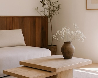 May include: A light-coloured, rectangular coffee table with a lower, smaller table beneath it. A brown vase with white flowers sits on top. A small tree in a pot and a sofa are in the background.