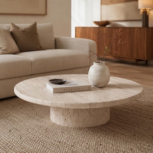 Op de afbeelding: Een ronde, lage salontafel gemaakt van beige, poreuze travertijnsteen met een cilindrische voet. Op het tafelblad liggen een wit boek met de titel KINFOLK, een kleine donkere kom en een ronde, gebroken witte keramische vaas met dunne takken.