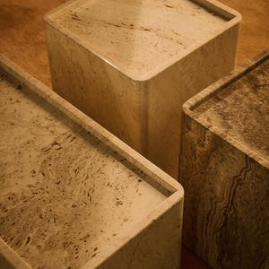 Op de afbeelding: Drie vierkante, beige stenen sokkels met afgeronde hoeken. De sokkels hebben een gemarmerd uiterlijk met bruine en bruingele aderen. De sokkels zijn geplaatst op een houten oppervlak.