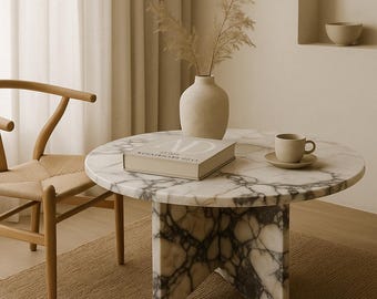 May include: A round marble coffee table with a white and gray veined pattern. A beige book, a cup and saucer, and a vase with dried plants are on the table. A wooden chair is to the left.