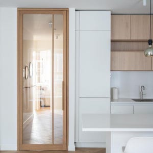 Op de afbeelding: Een modern keukeninrichting met een glazen deur met licht houten frame, witte kasten en bovenkasten van licht hout. De ruimte heeft een minimalistisch design met een wit aanrechtblad en een hanglamp.