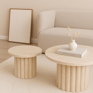 May include: Two light-colored, round coffee tables with fluted bases sit on a cream-colored rug. One table has a book titled "Yves Klein" and a small white vase with dried flowers. A framed abstract art piece and a beige sofa are in the background.