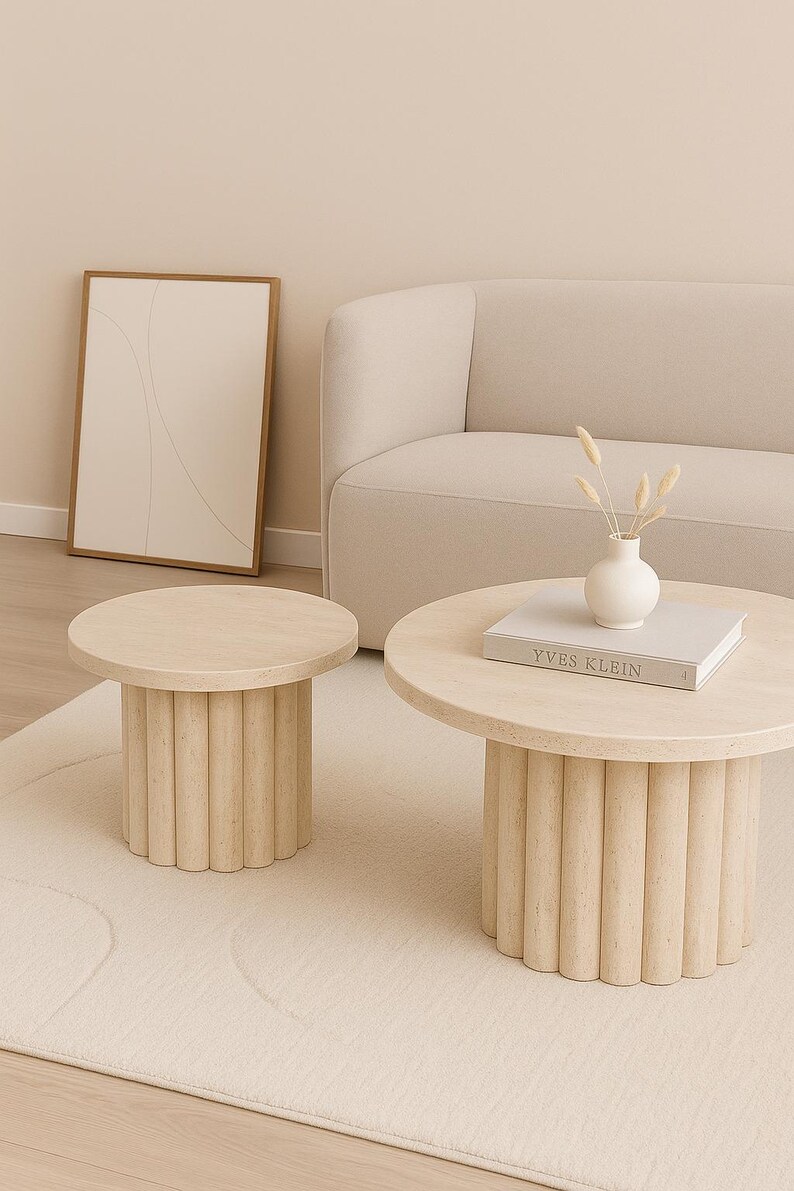 May include: Two light-colored, round coffee tables with fluted bases sit on a cream-colored rug. One table has a book titled "Yves Klein" and a small white vase with dried flowers. A framed abstract art piece and a beige sofa are in the background.