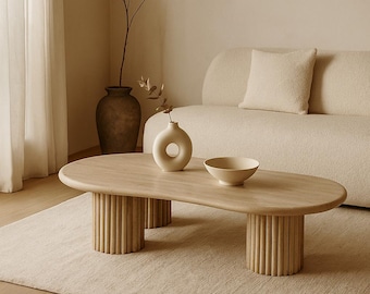 May include: Oval wooden coffee table with cylindrical legs. A small, round vase and a bowl sit on the table. The table is on a light-coloured rug, with a beige sofa in the background.