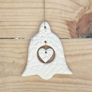 May include: A white ceramic bell-shaped ornament with a heart cutout and a smaller heart inside. The ornament has a textured surface and is suspended by a gold string. The background is a light brown wooden surface.