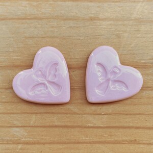 Puede incluir: Dos adornos de cer&aacute;mica en forma de coraz&oacute;n rosa claro. Cada coraz&oacute;n presenta un dise&ntilde;o en relieve de un &aacute;ngel. Los adornos se muestran sobre una superficie de madera marr&oacute;n claro.