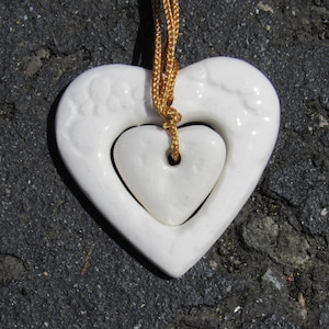 May include: A white ceramic heart pendant with a smaller heart inside, hanging from a gold cord.
