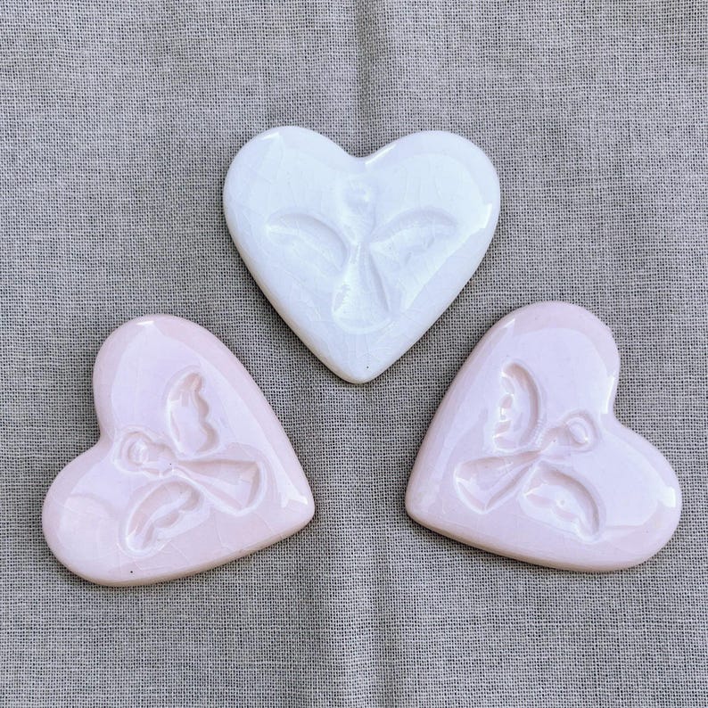 Puede incluir: Tres decoraciones de cer&aacute;mica en forma de coraz&oacute;n. Una es blanca con un dise&ntilde;o de rostro, y dos son rosas con un dise&ntilde;o de lazo. Los objetos est&aacute;n dispuestos sobre una tela gris.