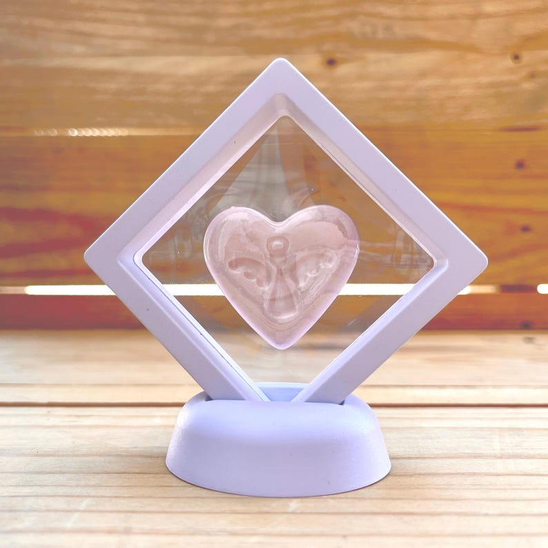 Puede incluir: Un expositor lila claro con una ventana transparente, que contiene un objeto en forma de coraz&oacute;n rosa. El coraz&oacute;n tiene un dise&ntilde;o de &aacute;ngel grabado. El expositor est&aacute; sobre una base lila claro, sobre un fondo de madera.