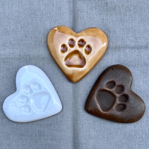 May include: Three heart-shaped ceramic ornaments with paw print designs. One is light brown with a darker paw print, another is white with a white paw print, and the third is a darker brown with a darker paw print.