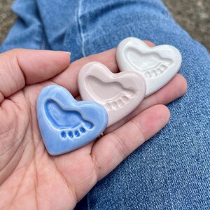 Puede incluir: Tres recuerdos de cer&aacute;mica en forma de coraz&oacute;n, cada uno con una huella de beb&eacute;. Los corazones son azul claro, rosa y blanco. Un regalo atento para un reci&eacute;n nacido o un baby shower.