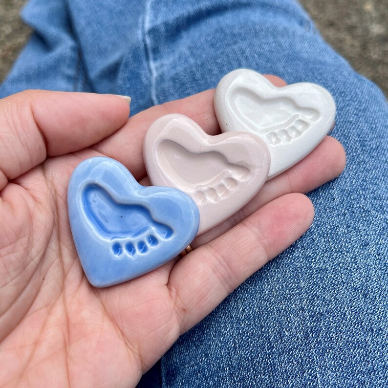 Puede incluir: Tres recuerdos de cer&aacute;mica en forma de coraz&oacute;n, cada uno con una huella de beb&eacute;. Los corazones son azul claro, rosa y blanco. Un regalo atento para un reci&eacute;n nacido o un baby shower.