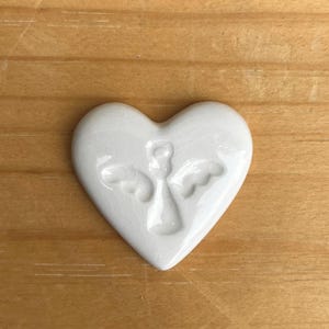 Puede incluir: Adorno de cer&aacute;mica blanca en forma de coraz&oacute;n con un dise&ntilde;o de &aacute;ngel en relieve. El &aacute;ngel tiene alas extendidas. El adorno est&aacute; sobre una superficie de madera.