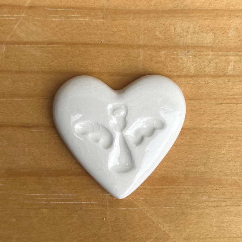 Puede incluir: Adorno de cer&aacute;mica blanca en forma de coraz&oacute;n con un dise&ntilde;o de &aacute;ngel en relieve. El &aacute;ngel tiene alas extendidas. El adorno est&aacute; sobre una superficie de madera.