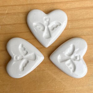 Puede incluir: Tres adornos blancos de cer&aacute;mica en forma de coraz&oacute;n con un dise&ntilde;o de &aacute;ngel en relieve. Los adornos est&aacute;n sobre una superficie de madera marr&oacute;n claro. Cada coraz&oacute;n tiene un acabado craquelado.