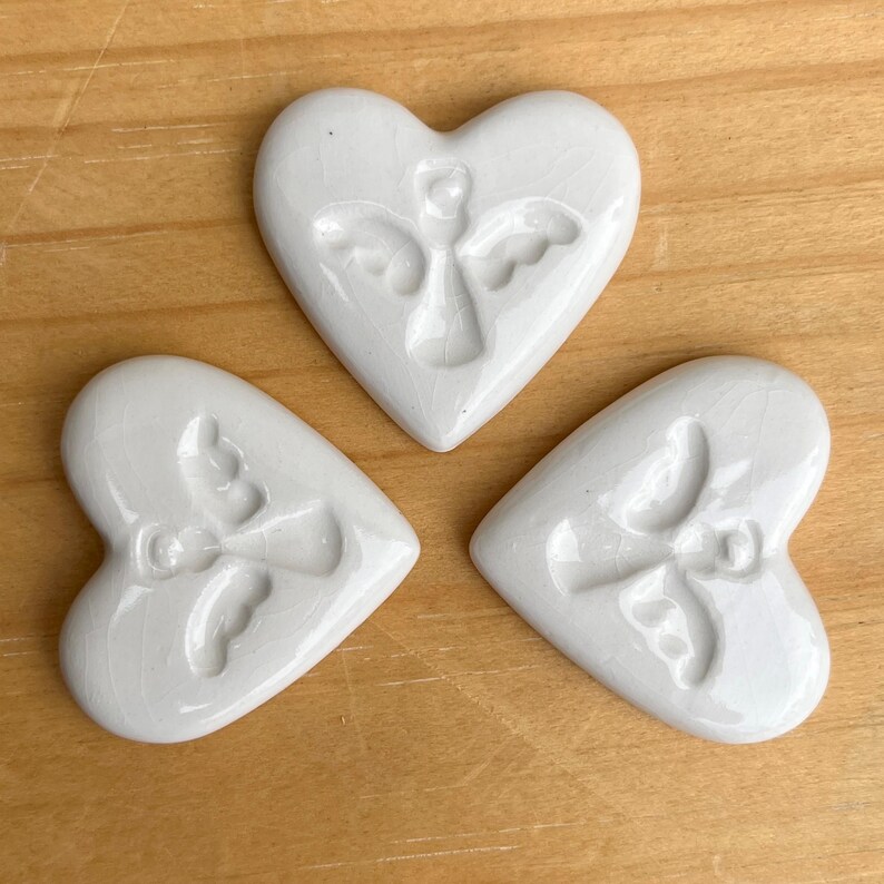 Puede incluir: Tres adornos blancos de cer&aacute;mica en forma de coraz&oacute;n con un dise&ntilde;o de &aacute;ngel en relieve. Los adornos est&aacute;n sobre una superficie de madera marr&oacute;n claro. Cada coraz&oacute;n tiene un acabado craquelado.