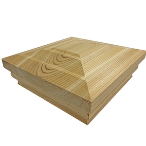 May include: A square, unfinished wooden post cap with a light brown color. The cap has a layered design with a beveled top and decorative molding along the bottom edge. The wood grain is visible.