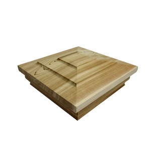 May include: A square wooden post cap with a layered, pyramid-shaped design. The cap is made of light brown wood with a natural grain pattern.