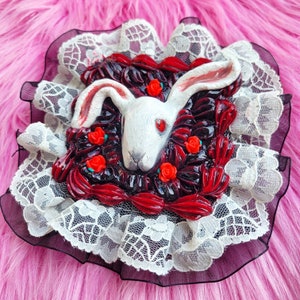 以下が含まれることがあります： 赤い目をした白いウサギの頭が、赤と黒のバラに囲まれています。ウサギの頭は、黒い裏地が付いた白いレースのトリムに付けられています。