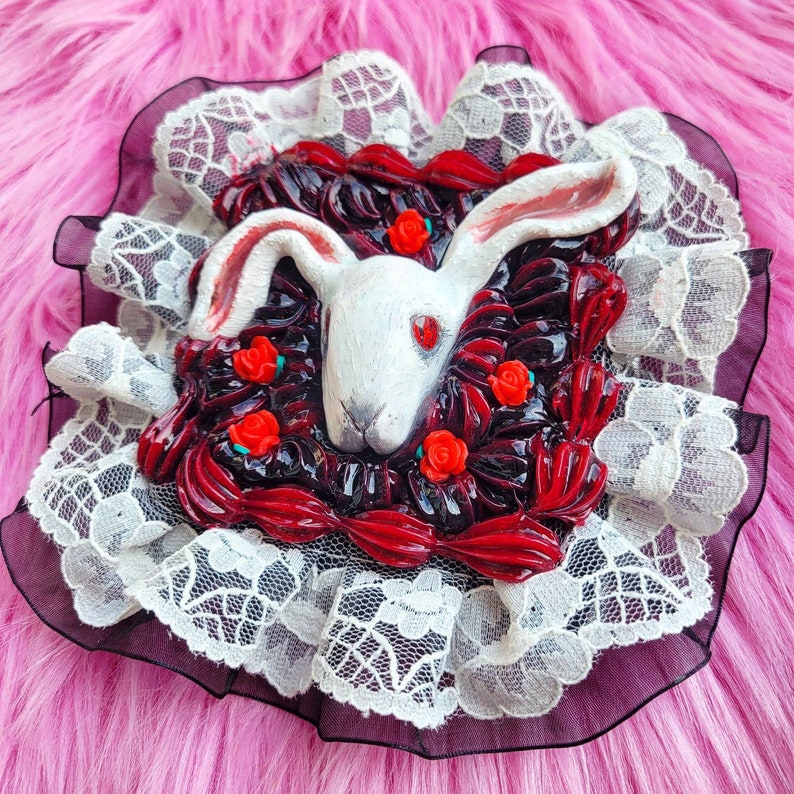 以下が含まれることがあります： 赤い目をした白いウサギの頭が、赤と黒のバラに囲まれています。ウサギの頭は、黒い裏地が付いた白いレースのトリムに付けられています。