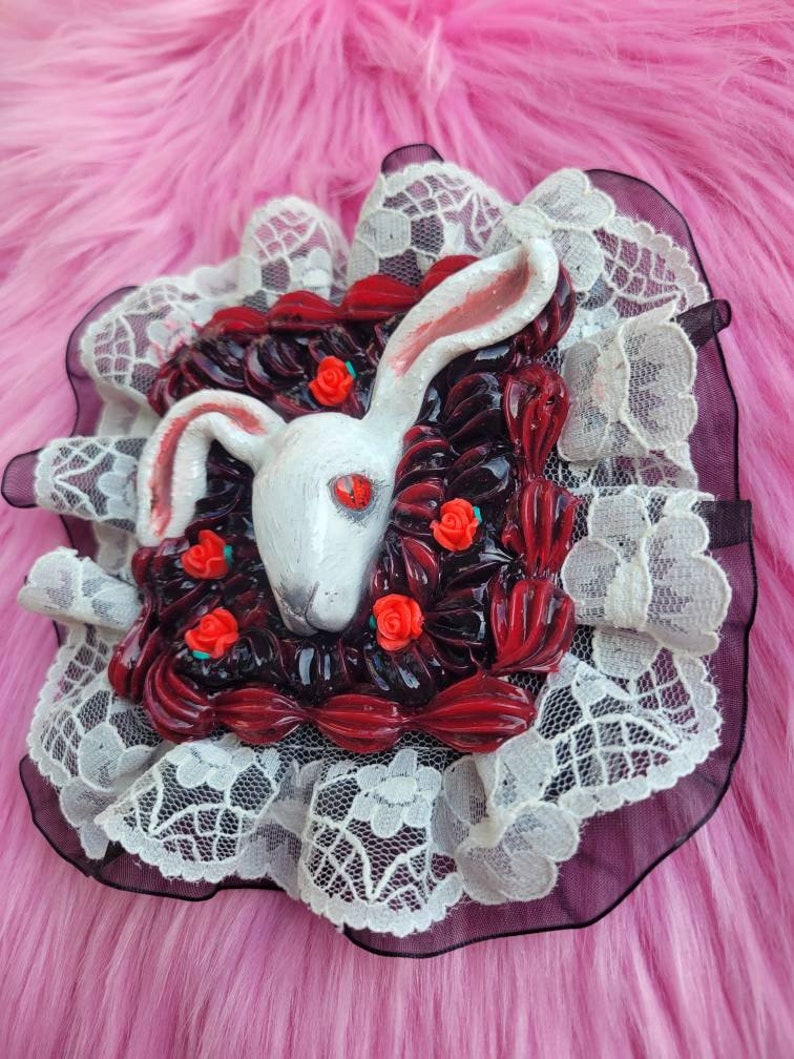 以下が含まれることがあります： 赤い目をした白いウサギが、赤いバラと黒い葉に囲まれています。ウサギは、白いレースの縁取りが施された正方形のフレームの中にいます。