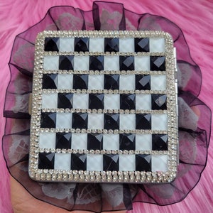 以下が含まれることがあります： 黒と白の市松模様の銀色のタバコケース。ケースはラインストーンで覆われています。