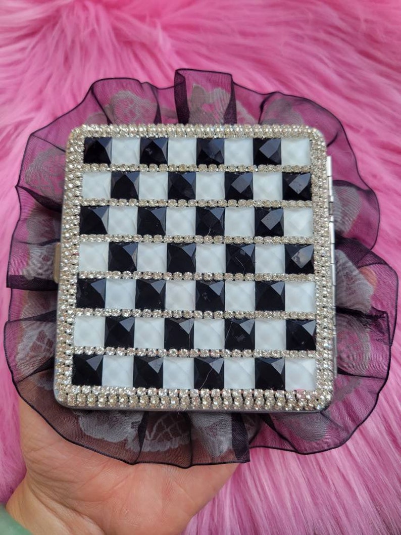 以下が含まれることがあります： 黒と白の市松模様の銀色のタバコケース。ケースはラインストーンで覆われています。