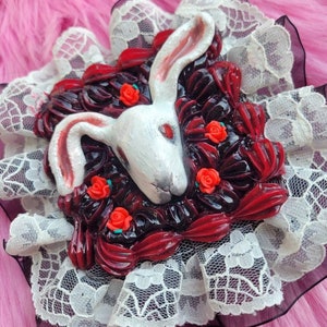 以下が含まれることがあります： 白いウサギの頭が、赤いバラと黒と赤の渦巻きで飾られ、白いレースで囲まれています