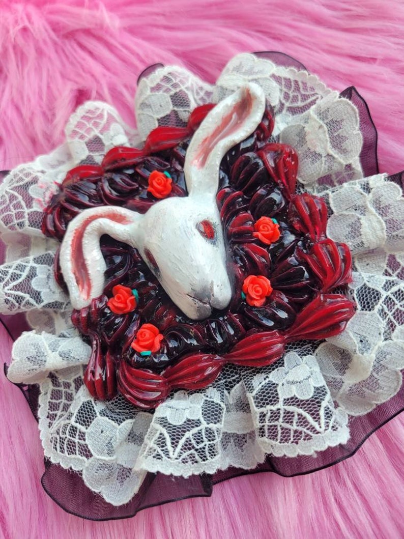以下が含まれることがあります： 白いウサギの頭が、赤いバラと黒と赤の渦巻きで飾られ、白いレースで囲まれています