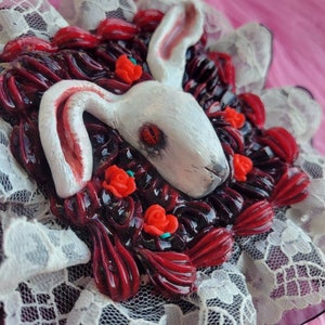以下が含まれることがあります： 赤い目をした白いウサギが、赤いバラと黒いレースで囲まれています。ウサギはポリマークレイで作られており、ブローチです。