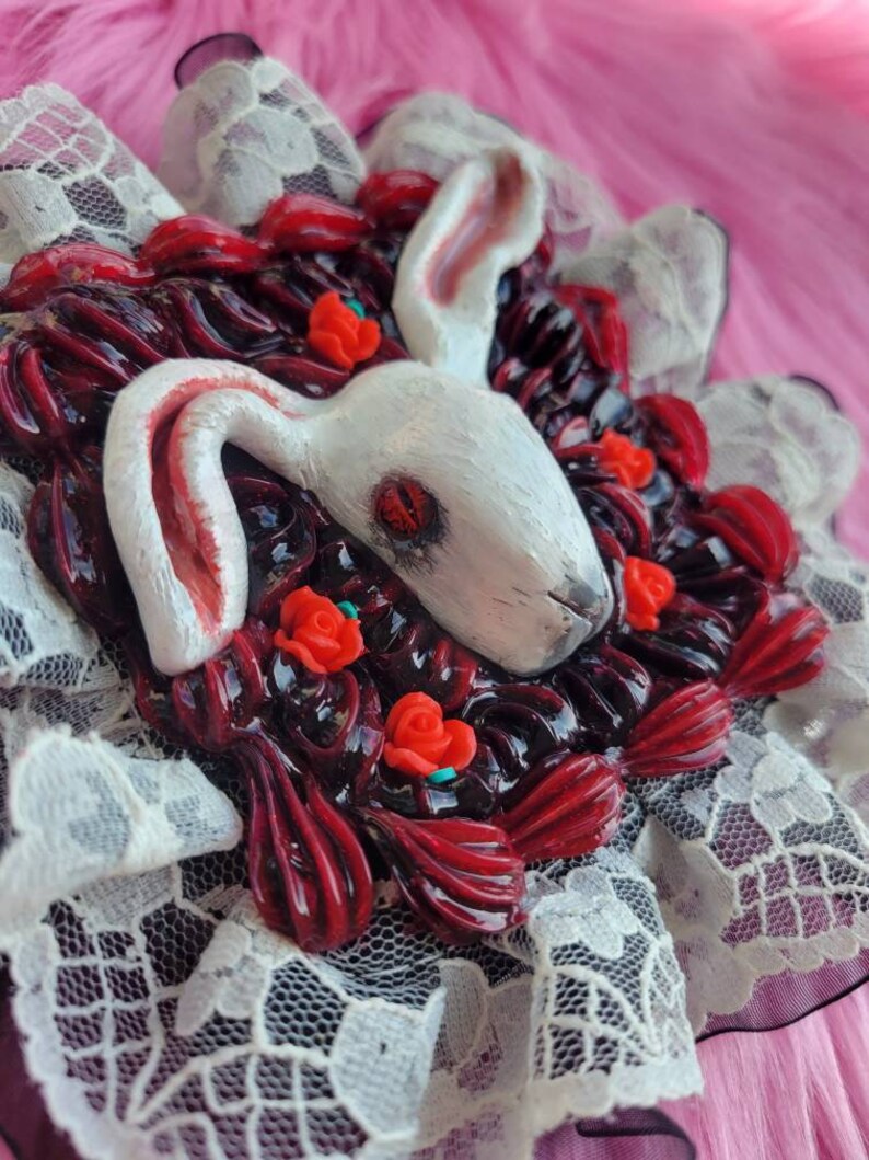以下が含まれることがあります： 赤い目をした白いウサギが、赤いバラと黒いレースで囲まれています。ウサギはポリマークレイで作られており、ブローチです。
