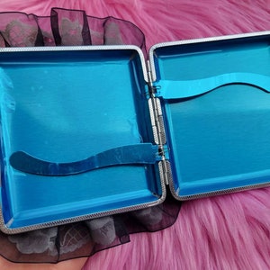 以下が含まれることがあります： 青い内側と黒いレースのトリムが付いた銀色のタバコケース。ケースが開いていて、内側が見えています。