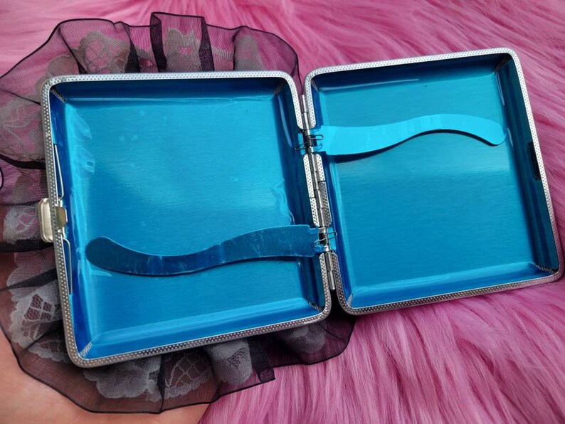 以下が含まれることがあります： 青い内側と黒いレースのトリムが付いた銀色のタバコケース。ケースが開いていて、内側が見えています。