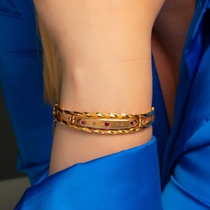 Pode incluir: Uma pulseira dourada com um design retangular central, adornada com pequenas pedras preciosas vermelhas e detalhes em forma de estrela. A pulseira apresenta um design trançado nas laterais e é usada no pulso.