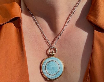 May include: A round, gold-toned pendant necklace with a light blue enamel face and a silver chain. The pendant has a detailed circular design in the center. The background is an orange-colored top.