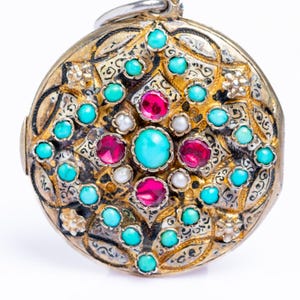 Puede incluir: Colgante redondo y ornamentado con una fianza plateada. El metal dorado está intrincadamente detallado con piedras preciosas turquesas, rojo rubí y blancas. El colgante tiene una estética vintage o antigua.