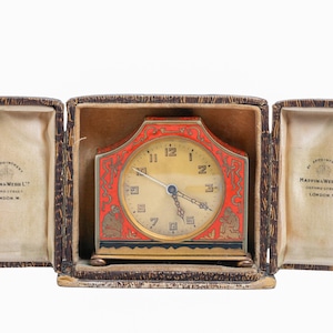 May include: An antique clock in a leather case. The clock face is cream-colored with black numerals and hands. The red frame has decorative gold accents. The case is open, revealing the clock and the interior lining with text.